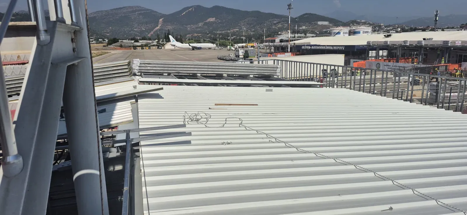 Toiture à nervures de teinte claire en cours de pose, avec des paquets de panneaux à gauche ; à l'arrière-plan, un avion sur l'aire de stationnement, le bâtiment du terminal et des collines